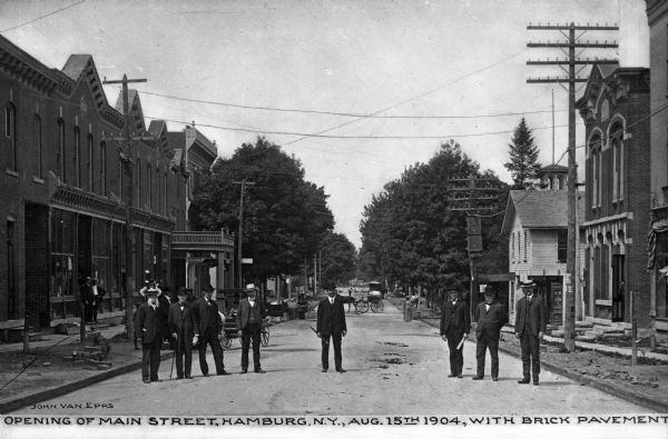 Men in suits and hats stand in the middle of the newly paved main street to pose for the commemorative photograph. The street is lined with brick buildings and horse-drawn carriages are in the distance.  Caption reads, "Opening of Main Street, Hamburg, N.Y., Aug. 15th 1904, with brick pavement."