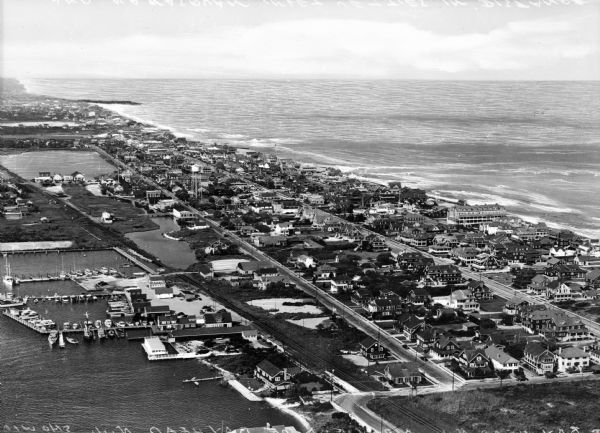 Aerial view of a developed strip of land on Barnegat Bay showing the Atlantic ocean, houses, lakes, railroad tracks, a marina and a jetty in the distance.