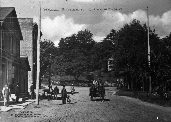 Street level view of a dirt road through a country town with pedestrians looking on, a horse-drawn carriage approaching and the facades of a few brick and wooden buildings. Caption reads, "Wall Street, Oxford, N.J."