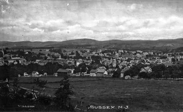 Distant view of a country town from across a pasture with rolling hills in the background. Caption reads, "Sussex, N.J."