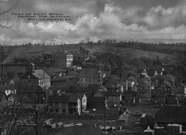 Elevated view of the West Ward of town including the hospital. View features many large wooden buildings built on a hillside with a lumber yard in the foreground and a tree-dotted hillside in the background. Caption reads, "Part of West Ward. Showing the Hospital, Belleponte, PA."