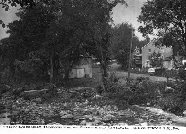 View looking north from a covered bridge, including a rocky stream-bed and a tree-lined dirt road with houses and other wooden buildings. Caption reads, "View looking north from covered bridge, Zieglerville, PA."