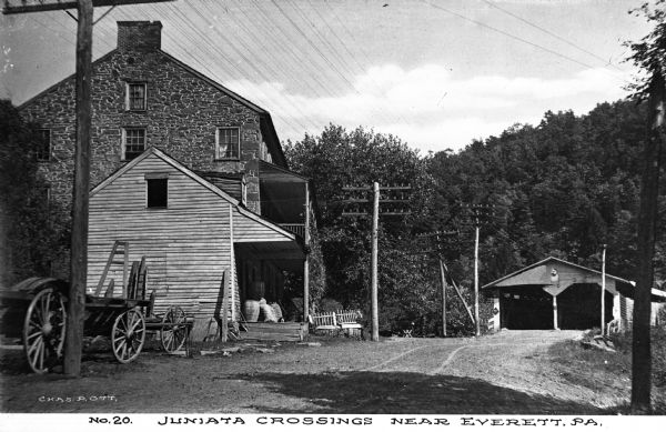 Juanita Crossing from a dirt road including a covered bridge, a stone farmhouse with a wooden addition, empty wagons and surrounding woods. Caption reads, "Juniata Crossings Near Everett, PA."