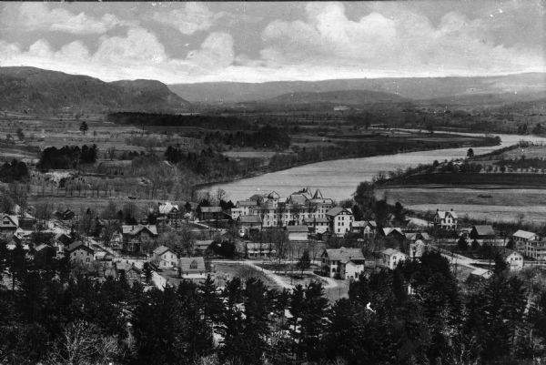 Elevated view of country town showing large. There are wooden buildings alongside the Delaware River which curves through fields and patches of woods in the distance. The Pocono mountains rise in the background.