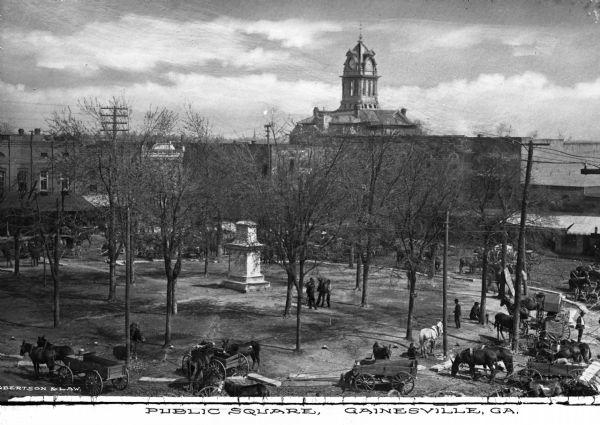 Elevated view of the town square with a stone monument and trees ringed by horse-drawn wagons and tall buildings. In the background an elaborate clock tower rises above the roof-lines. Caption reads, "Public Square, Gainesville, GA."