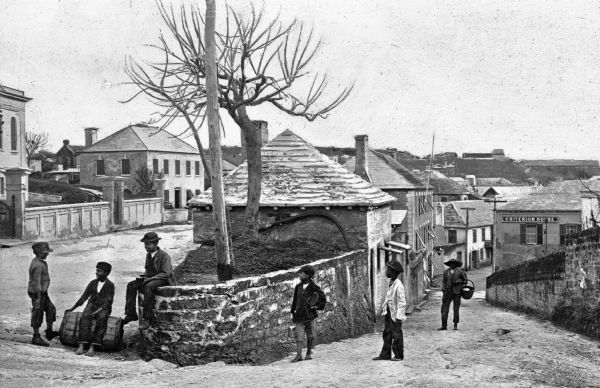 The apex of two streets in a densely built town in Bermuda. Five boys relax by a stone wall and small tree, and rooftops stretch into the distance down the hill. There is a man carrying a basket standing in the background. A building in the background on the right has a sign that reads: "Criterion House."