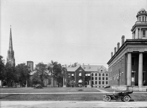 View of a grassy park bordered by four large stone and brick buildings.  A convertible is parked on the cobbled street and the the buildings all have different architectural styles.