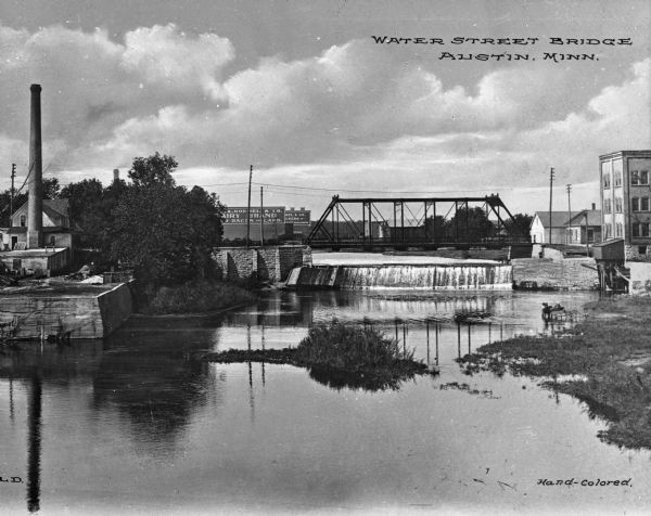 Waterside view of the Red Cedar River spanned by a metal bridge. The river has a small waterfall created by a dam and a horse-drawn carriage is standing in the shallows. In the background the Hormel factory is visible and other buildings also line the shore. Caption reads, "Water Street Bridge, Austin, Minn."