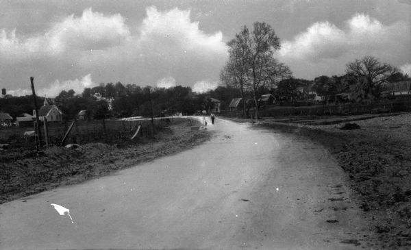 Low perspective of a dirt country road curving into the distance.  The road is bordered by fields and scattered wooden buildings while a solitary figure approaches from the distance.