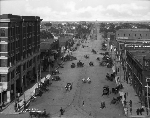 Elevated view of East Broadway, a wide road in the business district.  The street has tall stone and brick buildings as well as broad sidewalks. Many horse-drawn carriages, wagons and one car are on the street while a few pedestrians traverse the sidewalks. Streetcar tracks and cables are visible in the middle of the road.
