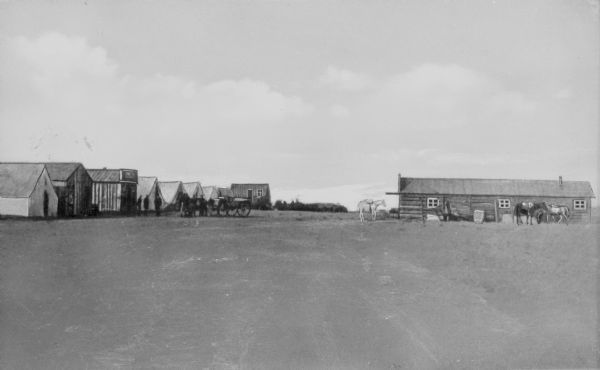 Distant view of the fledgling town showing a few log cabins, large tent buildings, saddled horses, pedestrians and a horse-drawn wagon.