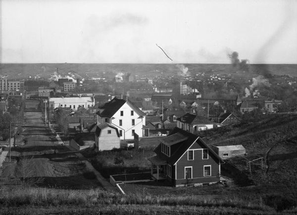 Elevated distant view of town from the top of a steep street that dead-ends at a field. In the foreground, large houses sit in ample yards while, in the background is a more dense settlement with tall buildings and trails of chimney smoke.