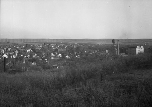 Distant view of town from a bracken covered hillside. A water tower rises above the rural town and a railroad bridge stretches across the background.