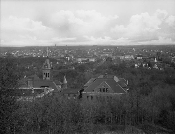High and distant view of city with a mansion's rooftop in the foreground. Young, close-growing leafless trees surround the mansion and extend to the edge of the city.