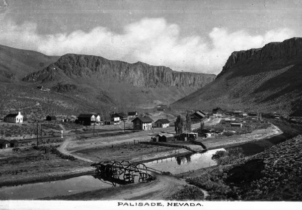 Elevated view of a small settlement between two mountains and alongside a river with tracks of the first transcontinental railroad passing through. A bridge spans the river. Caption reads: "Palisade, Nevada."