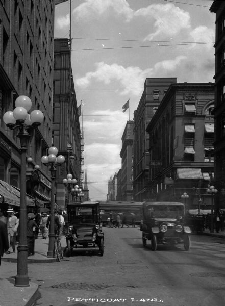Urban street view including cars, pedestrians and streetcar. Caption reads: "Pettitcoat Lane."