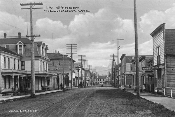 A view of First Street, with houses and shop fronts. Residences and storefronts line the unpaved road and utility lines run along both sides of the street. Down the street on the left is the Allen House, and on the right are signs for a Rooming House, Steam Dye Works and a Bakery. Caption reads: "1st Street, Tillamook, Ore."
