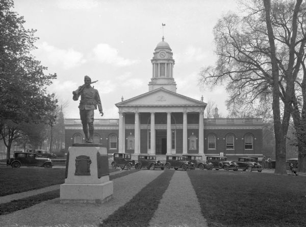A view of the the front facade of the town hall and of the World War I memorial statue. A line of cars are parked in front of the building.