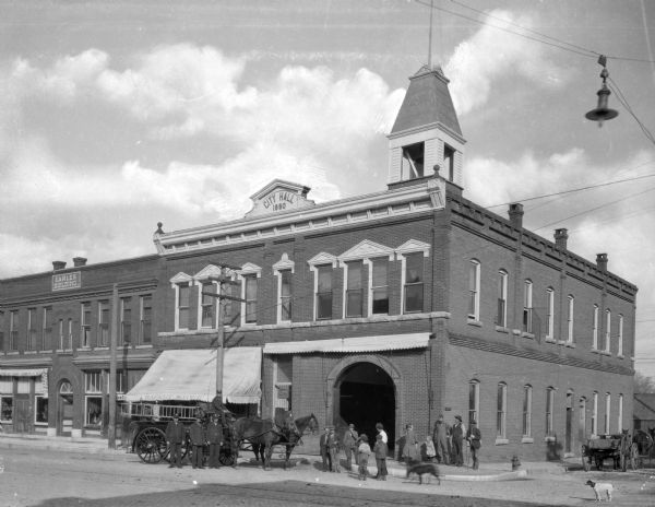 A view of City Hall and the fire department. A horse-drawn fire engine with fire fighters and dogs standing in front of the entrance to the fire station garage.