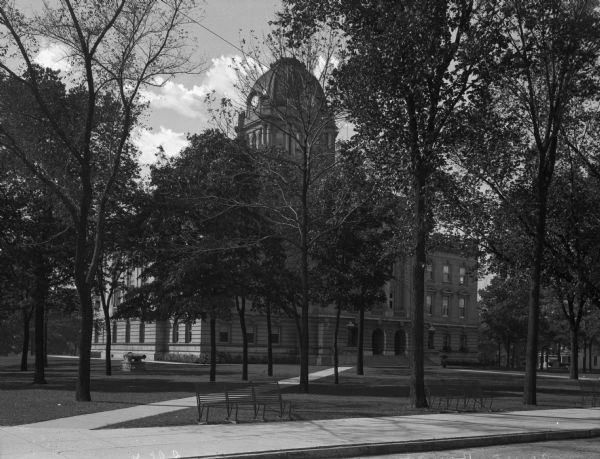 A view of the the courthouse with a clock tower. The building is somewhat obscured by trees, and there are benches near the sidewalk.