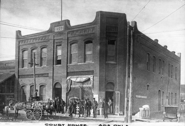 A view of the front and side of the courthouse. Men are gathered on the sidewalk. Published by the Sprague Brothers.