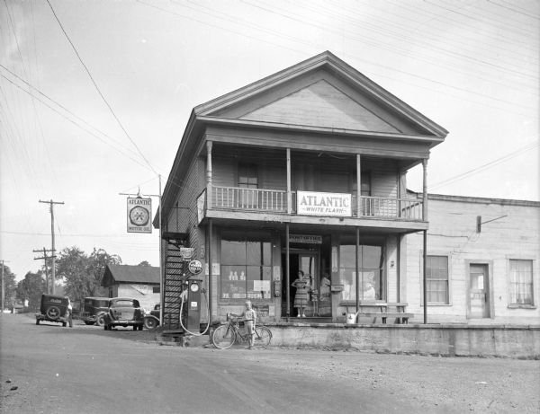 View down road toward the post office and gas station. A woman stands in front of the doorway, a young boy stands with a bicycle next to the gas pump, and automobiles are parked towards the rear of the building. A sign on the railing of the balcony reads: "Atlantic White Flash."