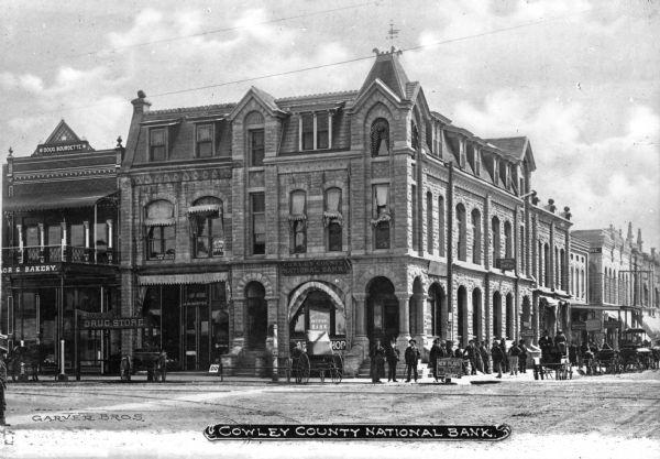 View across intersection toward the Cowley County National Bank. Other buildings and businesses are on either side, including a Drug Store and Bakery, and men stand on the corner in front of the bank. Caption reads: "Cowley County National Bank."