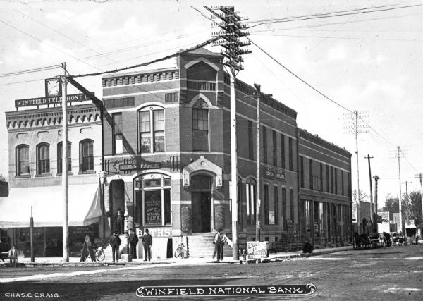 View across intersection toward the Winfield National Bank and surrounding businesses. Men stand on the sidewalk. Caption reads: "Winfield National Bank."