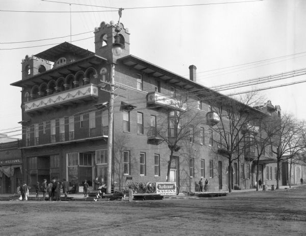 View across intersection toward the Baldwin Hotel and surrounding shops.  Pedestrians are on the sidewalk and in the street. A post office is on the corner. A sign on the sidewalk on the right reads: "The Strong, Durable & Light Running 'Old Hickory' Wagons, Sold By D.W. Brown Co." The sign is surrounded by wagon wheels.