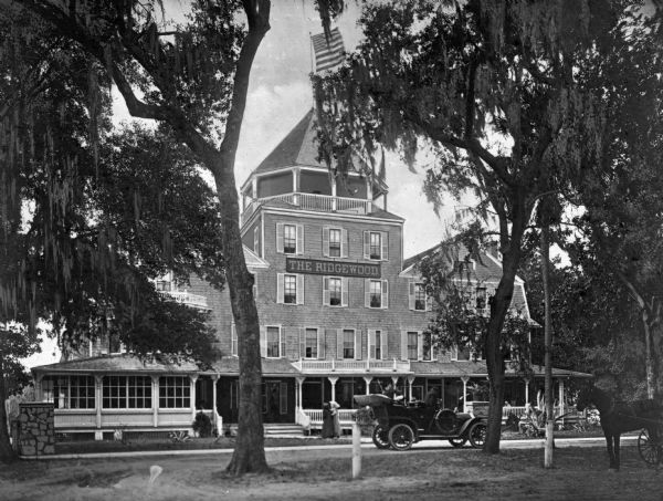 View across lawn toward the Ridgewood Hotel with an automobile parked in front. A flag is flying from the top of the hotel. A horse-drawn vehicle is on the right.
