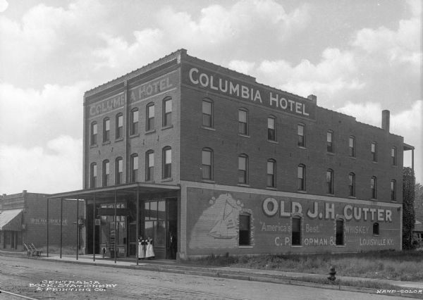 View across street toward the Columbia Hotel. Two women stand in front of the hotel under the roofed entrance. The side of the hotel has a painted advertisement for "Old J. H. Cutter Whiskey."