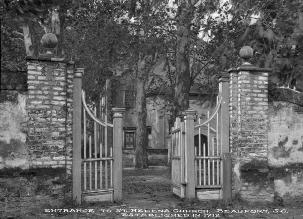 View of the stone wall and gate to St. Helena Episcopal Church. Trees partially conceal the building. Caption reads: "Entrance to St. Helena Church, Beaufort, S.C. Established in 1712."