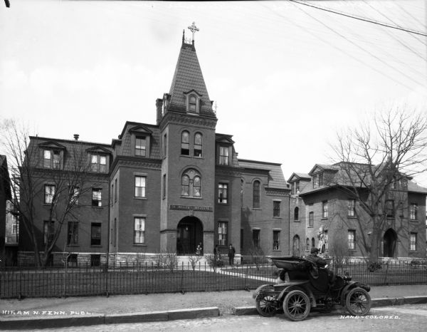 A view of the entrance and front facade of St. Joseph's Hospital. A man stands on the sidewalk near the entrance. A fence surrounds the building, and a man is sitting in a cars parked in front.