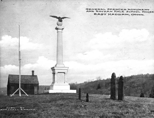 View of the Nathan Hale Schoolhouse, with the General Spencer Monument in the foreground. Erected by the State on the petition of the Nathan Hale Memorial Chapter of the Daughters of the American Revolution. General Spencer's body and that of his wife, as well as their original headstones, were later removed from their first resting place and located near the monument. Four grave stones are in the foreground and hills are in the background. Caption reads: "General Spencer Monument and Nathan Hale School House, East Haddam, Conn."