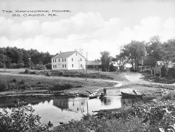 View over stream toward the Hawthorne House, childhood home of American novelist and short story writer Nathaniel Hawthorne (7/4/1804 – 5/19/1864) built around 1812. Canoes are docked by the edge of the water, and other homes are in the background. Caption reads: "The Hawthorne House, So. Casco, Me."