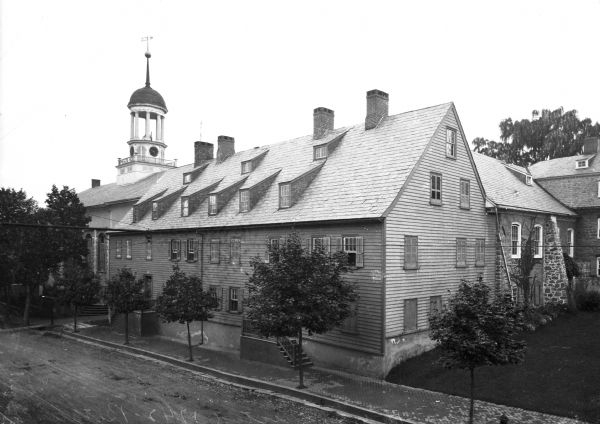 A view taken above street level of the Gemein House, which was built in 1742.  Trees line the street.  Other buildings appear beside and behind the house.    A telephone or electric line runs across the middle of the photograph.