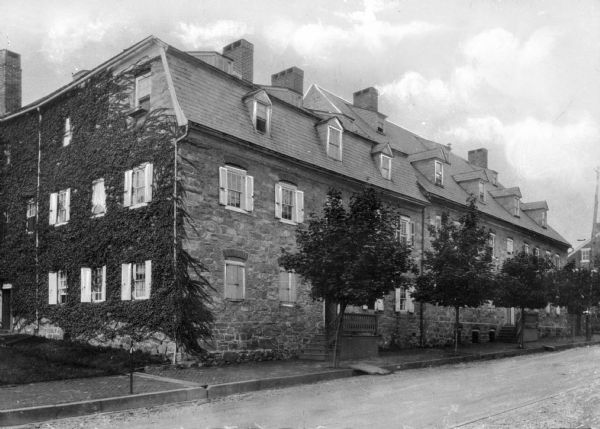 View across street toward the Moravian Sisters House. Vines cover the left side of the house, while trees lining the street slightly obscure the facade.