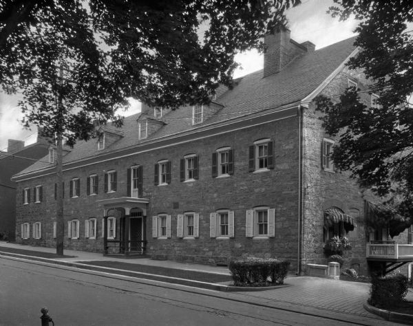 View across street toward the Widows House, built 1768. The trees lining the street slightly obscure the roof and right side of the house. The bottom window on the right side has an awning and a window box for flowers.