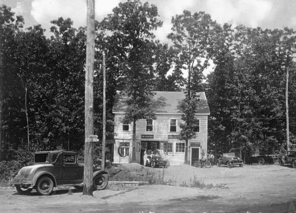 View toward the Community Hall in Sound Beach, Long Island, New York, serving as the Fire Department, Highway Department, and Police Department. Numerous men, automobiles, and a motorcycle are in front of the building, which is surrounded by trees.