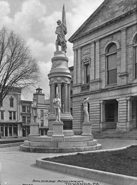 A view of the Soldiers' Monument in front of a stone building. Storefronts are in the background. Caption reads: "Soldiers' Monument Towanda, PA."