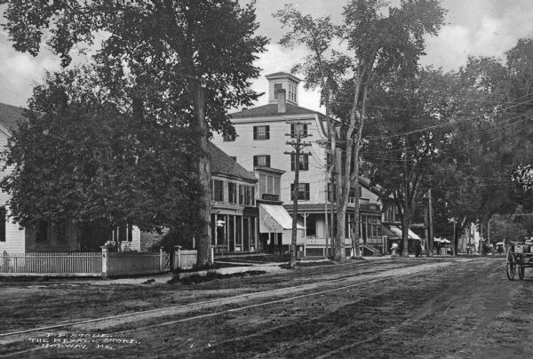 Angled view down unpaved road toward a tree-lined street and homes. Streetcar or train rails are along the left, and a cart is on the right. Caption reads: "