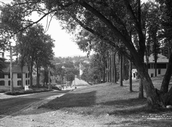 View along side of road lined with trees looking downhill toward residences on Cottage Street. Ladies in white dresses are walking on the sidewalk further down on the left.