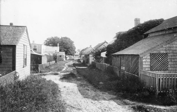 A view along the backs of cottages on a village lane in Siaconset, Nantucket Island. Shrubs line the unpaved road, and fences demarcate each cottage's yard.