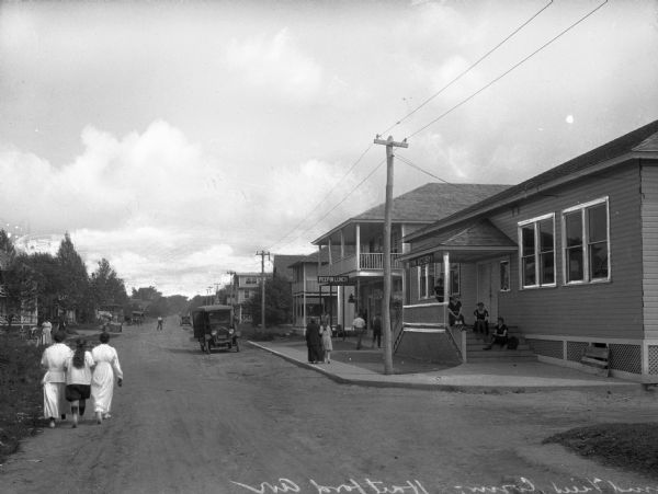 Slightly elevated view of Hartford Avenue. To the left is a field and trees, which partially conceals the buildings in the background, and to the right are storefronts. Men, women, and children are along the road and the sidewalks.
