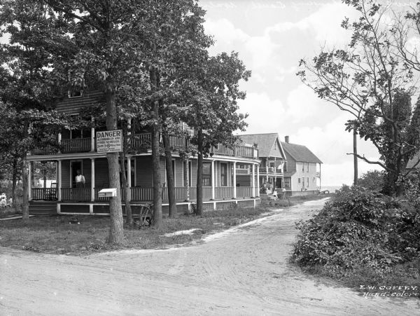 View from unpaved road toward houses down a side dirt road leading toward a beach in the background. A man stands on the porch of the house in the left foreground, and other people are on a porch further down. The sign on the tree in the foreground reads: "Danger, Automobiles and other Vehicles Slow to 6 miles per hour. Walnut Beach Improvement Association."