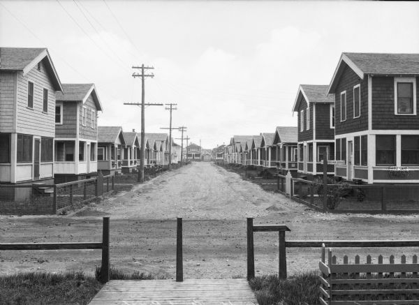 A street view of cottages, with a boardwalk and a fence in the foreground. Houses line both sides of the unpaved road.