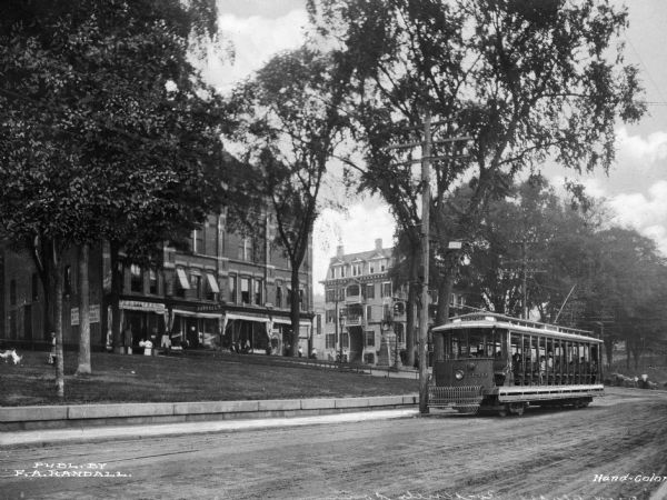 A street scene with a trolley car in the foreground. Up the small hill are two large buildings with people standing in front of the store windows, while a boy with two dogs play on the grassy hill.