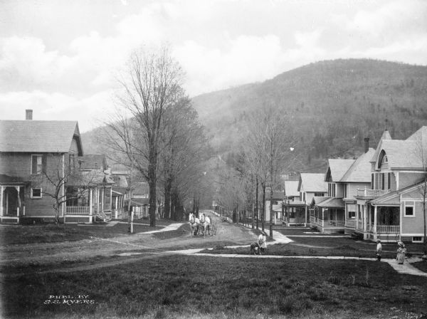 View across lawn toward the houses on tree-lined Walnut Street. A horse-drawn carriage is coming up the dirt street. A woman and children are on a sidewalk on the right.