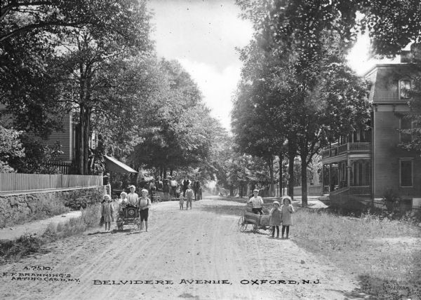 A view of Belvedere Avenue. The street is tree-lined, making the homes partially obscured. Men stand and pose in the middle ground, while three groups of children, boys and girls, pose in the front. Two of the groups of children have wagons. Printed in the bottom center: "Belvidere Avenue, Oxford, N.J."