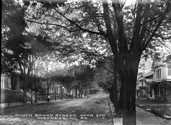 A view of S. Broad Street. Sidewalks line both sides of the streets.  Trees also line both sides, leaving most houses partially obscured. A few people can be seen in the foreground and middle ground. Caption reads: "South Broad Street, Near 3rd, Waynesboro, PA."